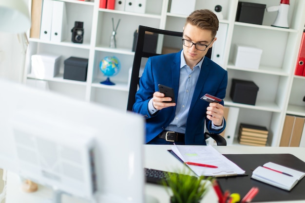 Ein junger Mann sitzt an einem Tisch im Büro und hält eine Bankkarte und ein Telefon.