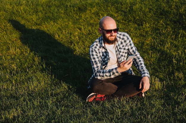 Ein junger Mann sitzt auf dem grünen Gras im Park und nutzt soziale Netzwerke mit einem Mobiltelefon