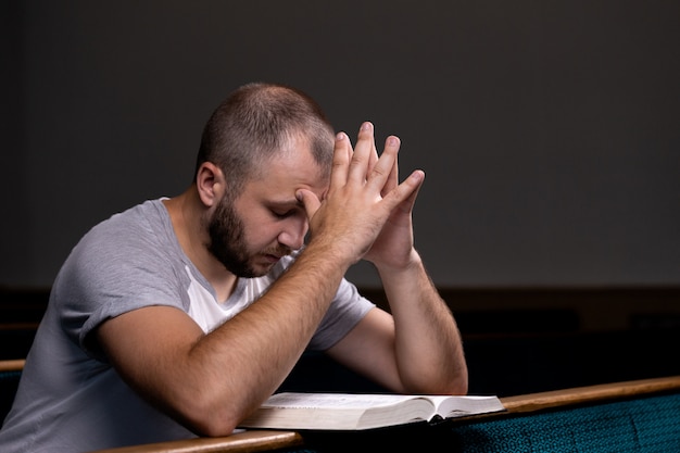 Ein junger Mann sitzt auf einer Kirchenbank, liest die Bibel und betet.