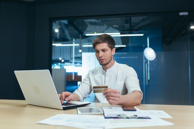 Ein junger Mann sitzt im Büro an seinem Schreibtisch und hält eine Kreditkarte in der Hand, die an einem Laptop arbeitet. Macht online Bestellungen und bezahlt Rechnungen