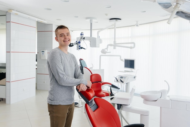 Ein junger Mann steht in der Nähe eines roten Behandlungsstuhls und lächelt in der modernen weißen Zahnheilkunde Behandlung und Vorbeugung von Karies bei Jugendlichen Moderne Zahnheilkunde und Prothetik