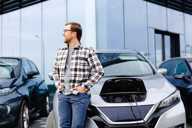 Ein junger Mann steht neben seinem neuen Elektroauto und ruht sich aus, während das Auto aufgeladen wird