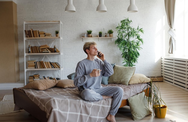 Ein junger Mann telefoniert mit einem Handy und sitzt mit einer Tasse Kaffee auf der Bettkante