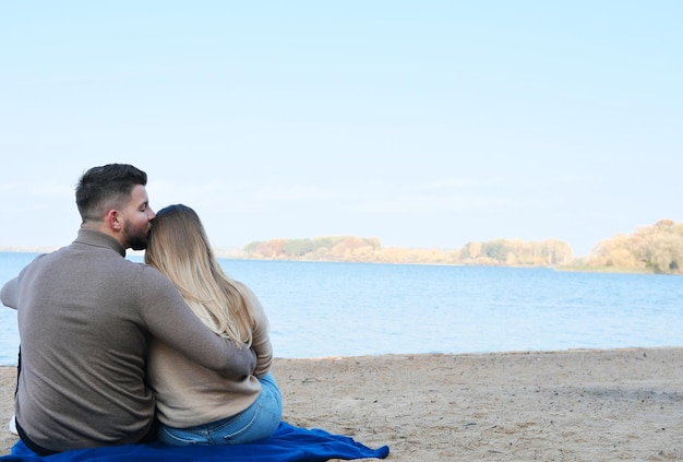 Ein junger Mann und eine junge Frau sitzen vor dem Hintergrund eines großen blauen Sees und umarmen sich Das Paar sitzt mit dem Rücken zur Kamera, der Typ küsst das Haar des Mädchens