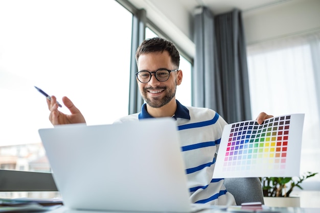 Ein junger Mann zeigt Farbproben auf dem Laptopbildschirm im Heimbüro und präsentiert ein Grafikdesign-Projekt bei einem Remote-Meeting. Ein Millennial-Designer wählt Gamma für die zukünftige Innenarchitektur des Hauses aus