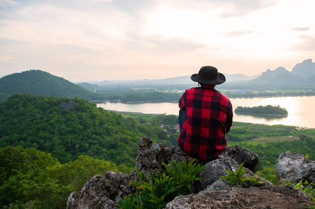 Ein junger Tourist in einem karierten Hemd und einem schwarzen Hut sitzt allein auf dem Berggipfel mit Blick auf die schöne Aussicht auf den See und den Sonnenuntergang. Er fühlte sich glücklich und entspannt, den Sonnenuntergang vom Berg aus zu beobachten