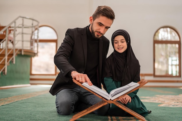 Ein junger Vater einer glücklichen muslimischen Familie mit einer kleinen Tochter, die ein heiliges Buch Koran in der Moschee liest