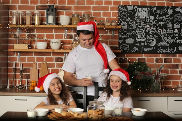 Ein junger Vater mit Weihnachtsmannmütze in der Küche steht neben seinen beiden Töchtern