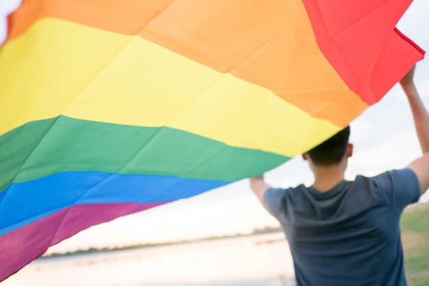 Ein junger weißer Mann sieht von hinten eine Regenbogenfahne über seinem Kopf halten.