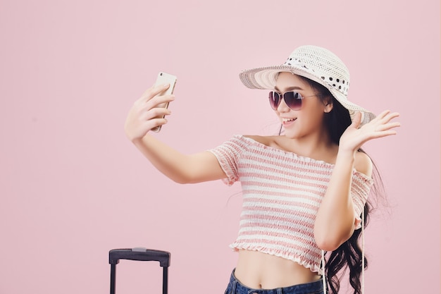 Ein junges asiatisches Mädchen mit einem hellen Gesicht, das einen Hut trägt und eine Brille trägt, macht ein Selfie im rosafarbenen Hintergrund des Studios. Pastellrosa-Tonfilter.