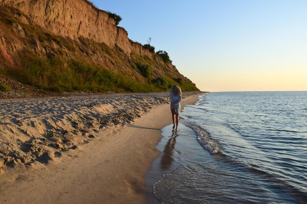 Ein junges blondes Mädchen geht barfuß am Strand entlang und trägt Schuhe in der Hand Rückansicht