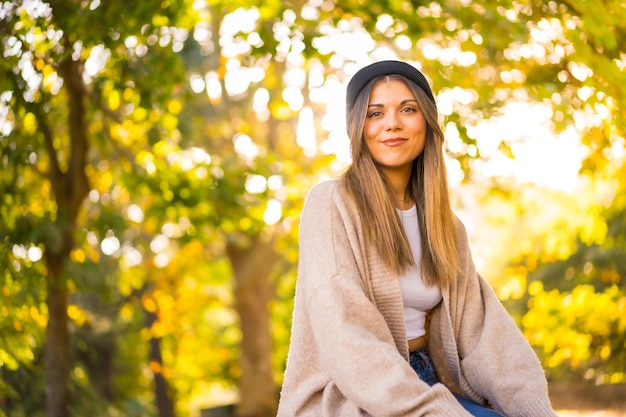 Ein junges blondes Mädchen in einem Wollhut im Herbst, das bei Sonnenuntergang im Park sitzt