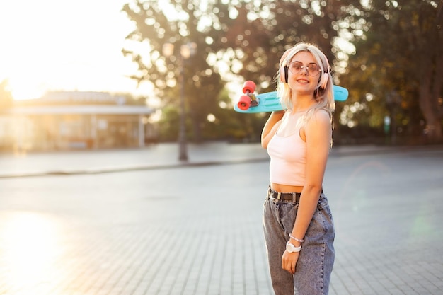 Ein junges cooles Hipster-Mädchen mit Kopfhörern und Sonnenbrille mit Skateboard verbringt Zeit in der Stadt