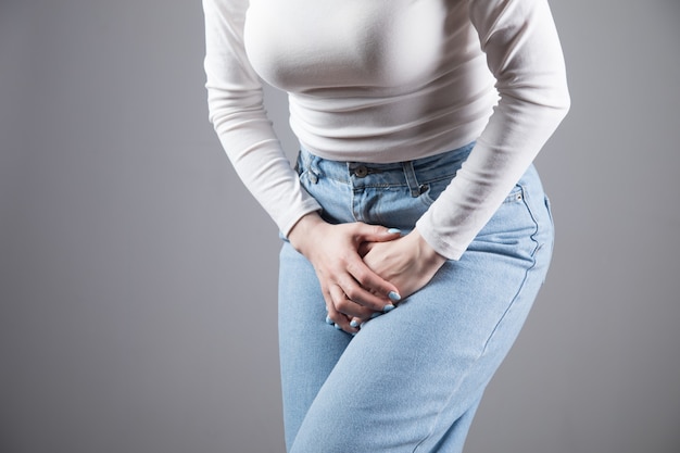 Ein junges dünnes Mädchen in Jeans hält ihre Hände zwischen ihren Beinen eingeklemmt. Frauengesundheit, Gynäkologie auf einer grauen Szene