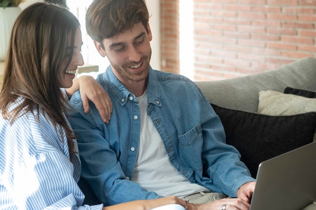 Ein junges Ehepaar sitzt zu Hause im Wohnzimmer auf dem Sofa und nutzt einen Tablet-PC für das Internet und soziale Medien. Eine glückliche Familie ruht sich auf der Couch aus, genießt das Wochenende, schaut sich Videos an oder spricht über einen Videoanruf.