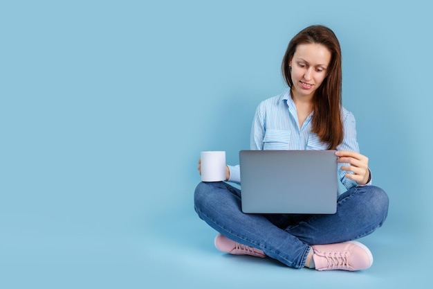 Ein junges erwachsenes Mädchen sitzt mit einem Laptop auf dem Schoß und hält eine weiße Tasse in den Händen, während sie in einen Laptop schaut. Blauer Hintergrund mit Exemplar