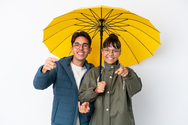 Ein junges gemischtes rassenpaar, das einen regenschirm isoliert auf weißem hintergrund hält, zeigt mit dem finger auf sie, während es lächelt