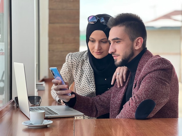 Ein junges glückliches muslimisches Paar, das zusammen an einem Laptop arbeitet und ein Mobiltelefon im Café-Restaurant oder Büro benutzt