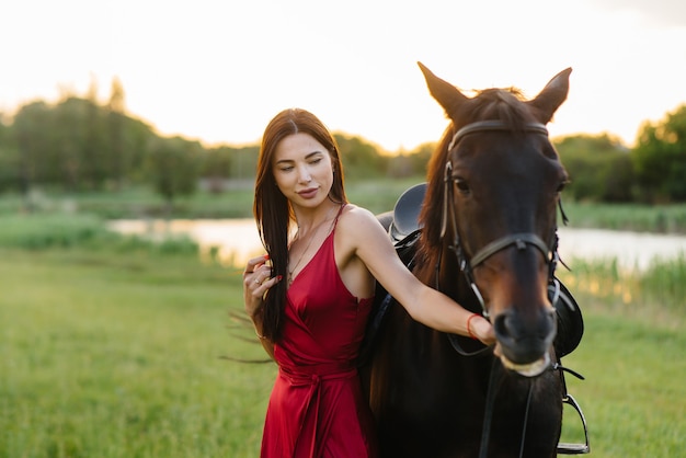 Ein junges hübsches Mädchen in einem roten Kleid posiert auf einer Ranch mit einem Vollbluthengst bei Sonnenuntergang. Liebe und Fürsorge für Tiere.