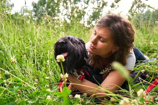 Ein junges hübsches Mädchen in einem weißen T-Shirt liegt auf dem Gras in der Natur mit einem schwarzen Mittelschnauzer-Hund