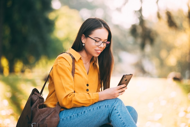 Ein junges hübsches Mädchen sitzt und sie schaut und lächelt in seinem Telefon draußen