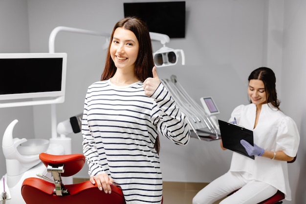 Ein junges hübsches Mädchen steht in der Nähe des Behandlungsstuhls im Büro und zeigt eine Fingerbewegung, während der Arzt Notizen über die Gesundheit des Patienten macht