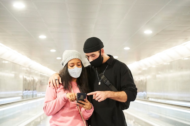 Ein junges interrassisches Liebespaar mit Masken und Wollmützen, das einen U-Bahn-Korridor entlang geht