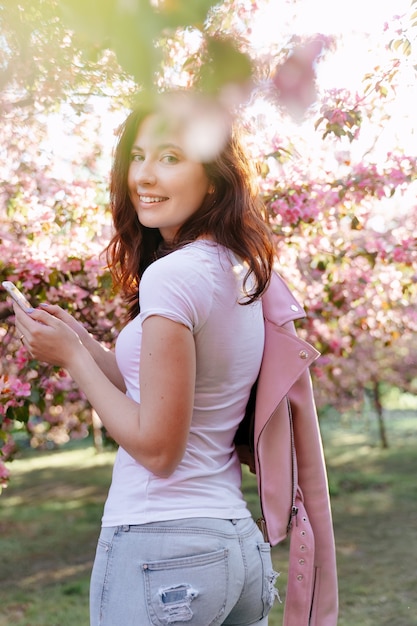 Ein junges lächelndes Mädchen steht mit einem Telefon in der Hand in einem blühenden Garten mit Apfel- und Kirschbäumen