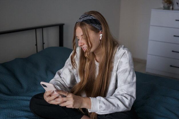 Ein junges Mädchen aus den 20ern mit einem Verband auf dem Kopf und langen blonden Haaren sitzt auf einem Bett in ihrem Wohnzimmer, hört Musik über Kopfhörer und schaut auf das Telefon