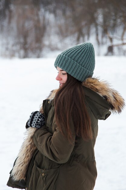 Ein junges Mädchen, brünett, in Pullover, Hut und grüner Jacke, vor dem Hintergrund der Winterlandschaft. Schnee und Frost, das Konzept von Weihnachten.