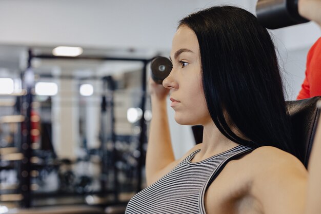 Ein junges Mädchen, das Sportkleidung in einer Turnhalle trägt, führt Hantelübungen durch, der Trainer hilft ihr