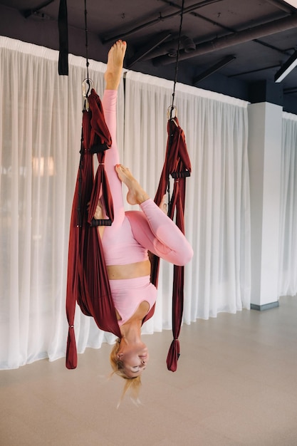 Ein junges Mädchen, ein Aero-Yoga-Trainer, hängt an einer Hängematte in der Yoga-Club-Halle, eine Frau in rosafarbener Sportkleidung macht Anti-Schwerkraft-Yoga