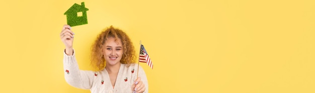 Ein junges Mädchen hält ein Modell eines grünen Hauses in ihren Händen und eine amerikanische Flagge Das Konzept des Kaufs eines Öko-Hauses