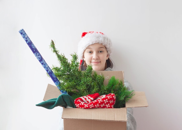 Ein junges Mädchen hält eine Schachtel mit einem künstlichen Weihnachtsbaum, einem Weihnachtspullover und einer Rolle Geschenkpapier hoch