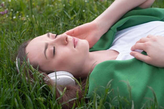 Ein junges Mädchen hört Musik über Kopfhörer, liegt auf dem grünen Gras im Park, lächelt und zeigt