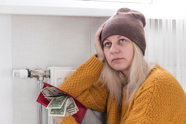 Foto ein junges mädchen in einem gelben pullover und einem braunen hut sitzt auf dem boden, zählt geld und denkt darüber nach, wie man rechnungen und steuern in der nähe einer heizung mit thermostat bezahlt