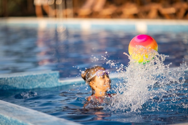 Ein junges Mädchen in einem hellen Leoparden-Badeanzug schwimmt an einem warmen, sonnigen Sommerabend mit einem bunten aufblasbaren Ball in einem tiefblauen Pool mit klarem, transparentem Wasser