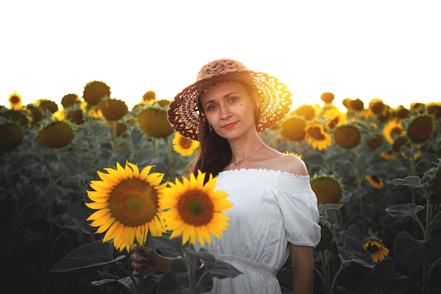 Ein junges Mädchen in einem weißen Kleid und Hut in einem Sonnenblumenfeld bei Sonnenuntergang Porträt einer Frau mit einer schlanken Figur auf einem Hintergrund aus gelben Blumen