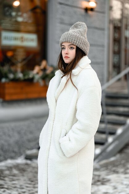 Ein junges Mädchen in einem weißen Pelzmantel und einem Hut steht im Winter vor einem Schaufenster auf der Straße