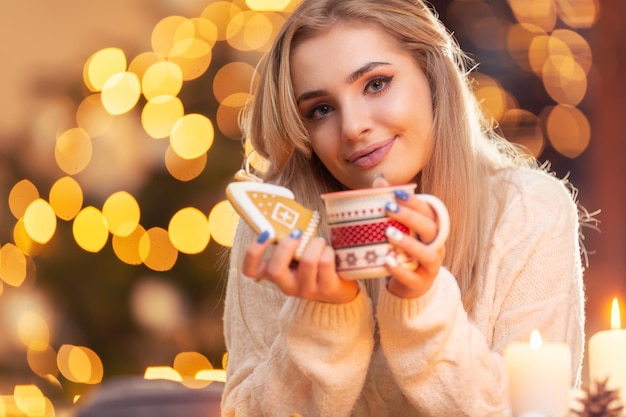 Ein junges Mädchen in einem Wollpullover, das einen Lebkuchen und eine Tasse mit Tee, Kaffee oder Weihnachtspunsch hält