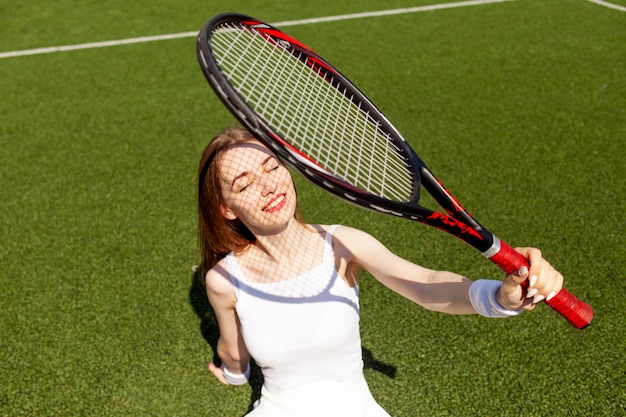 Ein junges Mädchen in einer weißen Sportuniform mit einem Tennisschläger sitzt und ruht auf einem grünen Platz