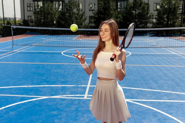 Ein junges Mädchen in einer weißen Uniform und mit einem Schläger wirft den Ball auf dem blauen Platz im Freien.