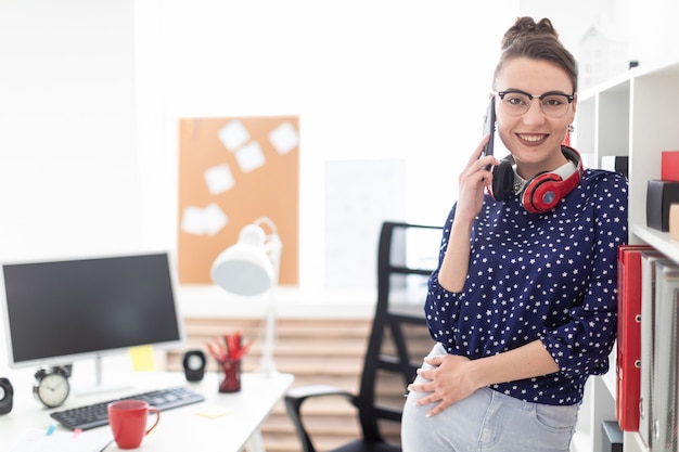 Ein junges Mädchen in Gläsern steht im Büro in der Nähe des Gestells und spricht am Telefon