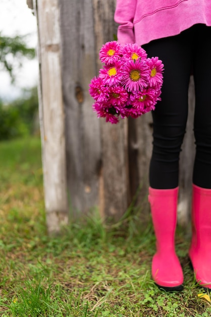 Ein junges Mädchen in rosa Gummistiefeln steht vor dem Hintergrund eines alten Hauses im Garten und hält einen Strauß rosa Blumen in den Händen