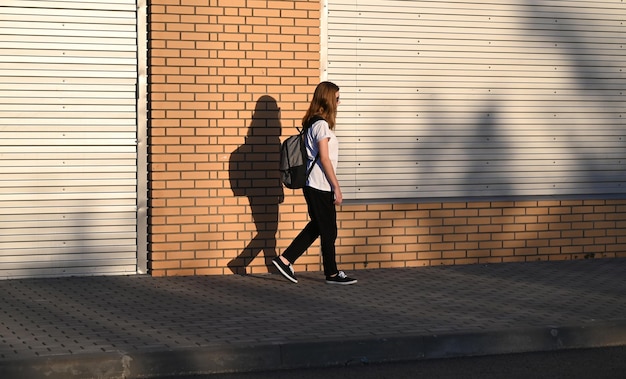 Ein junges Mädchen in schwarzer Hose und weißem T-Shirt geht mit einem schwarzen Rucksack auf dem Rücken die Straße entlang
