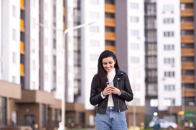 Ein junges Mädchen in schwarzer Lederjacke steht in einer Wohnanlage und schaut auf ihr Handy