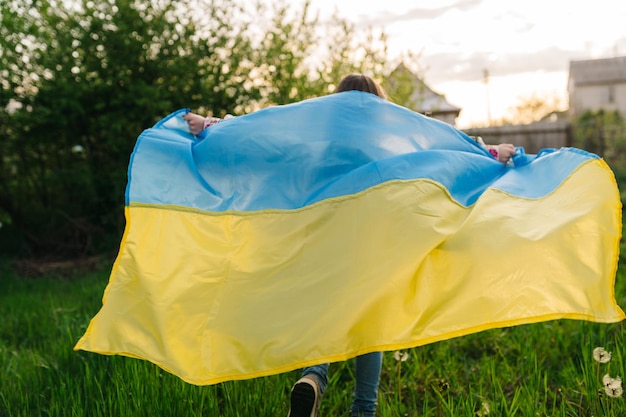 Ein junges Mädchen läuft mit der ukrainischen Flagge Foto eines Mädchens, das mit einer ukrainischen Flagge läuft, die als Regenmantel verwendet wird