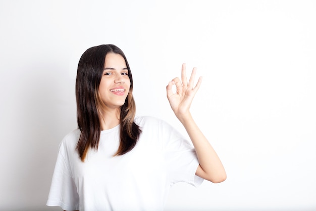 Ein junges Mädchen mit dunklen Haaren in einem weißen T-Shirt zeigt OK und lächelt Weißer Hintergrund