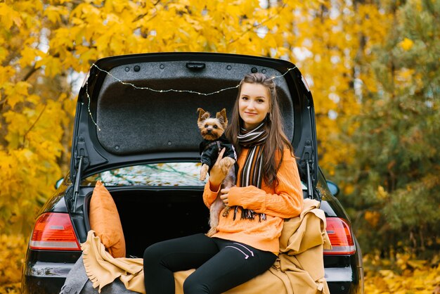 Ein junges mädchen mit einem hund sitzt im kofferraum eines autos vor dem hintergrund eines herbstwaldes. haustier und menschliche freundschaft