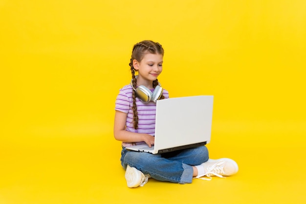 Ein junges Mädchen mit einem Laptop. Ein kleines Mädchen sitzt mit gekreuzten Beinen auf dem Boden und hält einen Laptop. Soziale Netzwerke für Teenager. Gelber, isolierter Hintergrund